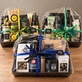 Tre gavekurve fra SMAGdansk med danske specialiteter, lakrids, chokolade, delikatesser og drikkevarer, pakket i cellofan med dekorative gavebånd.