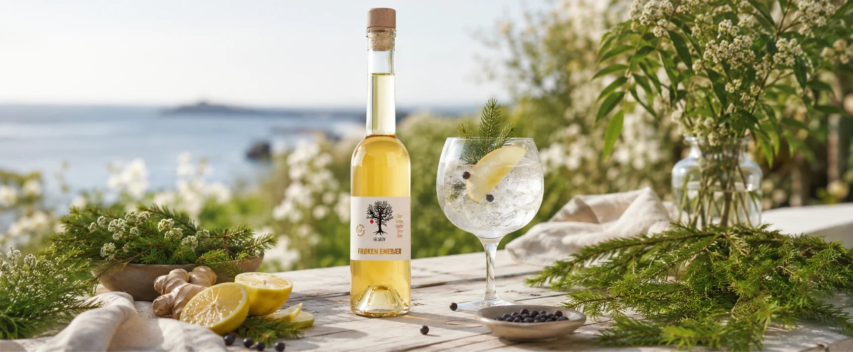 Hr. Skov Frøken Enebær serveret som frisk drink med is, æble og enebær i sommerlige omgivelser med grønne grene og udsigt over vandet.