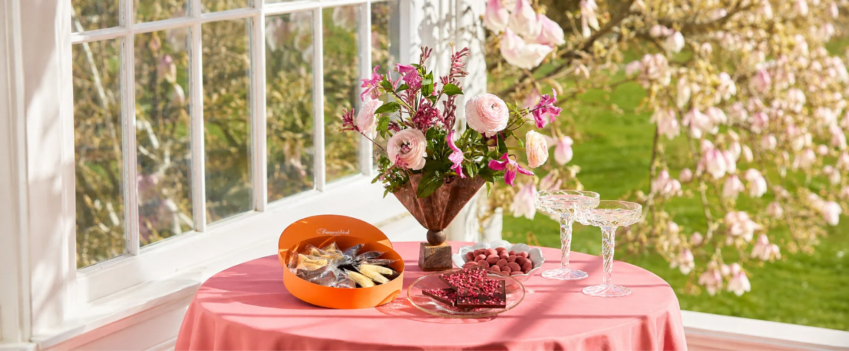 Summerbird chokolade og søde delikatesser serveret på et sommerligt bord med blomster, rosa nuancer og udsigt til en blomstrende have.