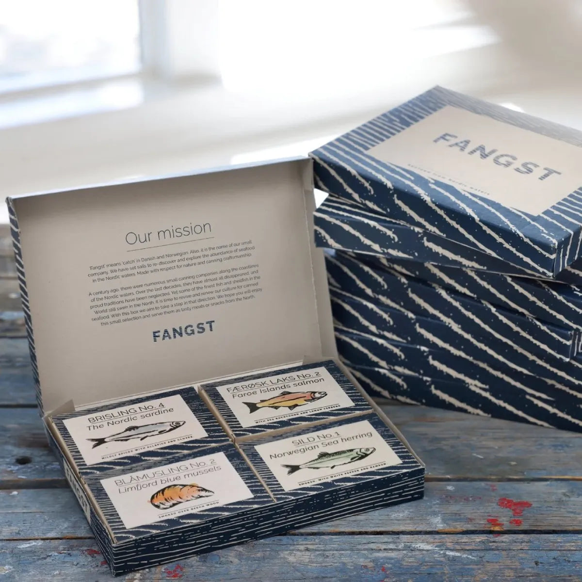 Fangst Giftbox gaveæske med 4 dåser nordisk seafood – udvalgte Fangst-varianter med sild, brisling, blåmusling og færøsk laks, perfekt som gave og til nordisk tapasbord. Her placeret på træbænk med uåbne gaveæsker til højre for sig.