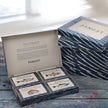 Fangst Giftbox gaveæske med 4 dåser nordisk seafood – udvalgte Fangst-varianter med sild, brisling, blåmusling og færøsk laks, perfekt som gave og til nordisk tapasbord. Her placeret på træbænk med uåbne gaveæsker til højre for sig.