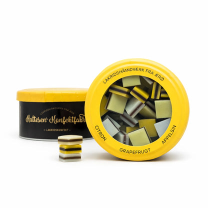 Præsentation af Hattesens Konfektfabrik Gul Æske 125 g – glutenfri og vegansk lakridskonfekt med citron, appelsin og grapefrugt, lakridshåndværk fra Ærø i elegant konfektæske. Her med en oprejst og en liggende æske. Desuden ses tre stykker konfekt fremme ved siden af æskerne.